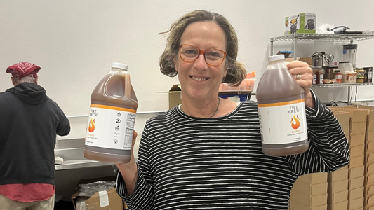 Smiling woman in striped shirt holds two large bottles of Fire Brew citrus tonic in a production room setting.Smiling woman in striped shirt holds two large bottles of Fire Brew citrus tonic in a production room setting.
