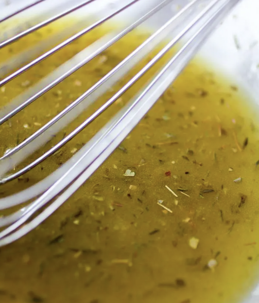 A close-up of a whisk resting in a bowl of herb-speckled vinaigrette, with visible oil and seasonings.