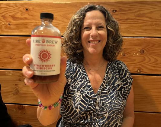 Smiling woman holding a bottle of Fire Brew Strawberry Hibiscus tonic while seated indoors against a wooden wall.