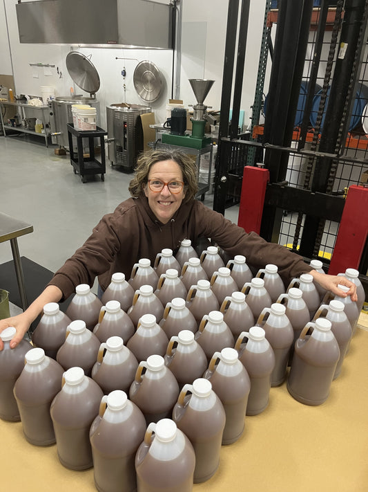 Smiling woman in a production facility with rows of large plastic jugs filled with brown liquid tonic.