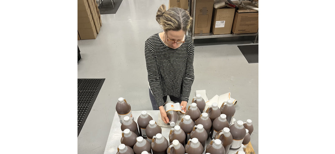 Woman labeling large brown bottles in a warehouse with boxes and packing supplies around.