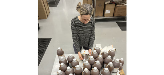 Woman labeling large brown bottles in a warehouse with boxes and packing supplies around.
