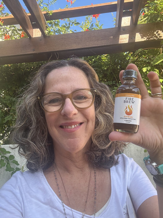 Smiling woman holding a small bottle of Fire Brew citrus tonic outdoors under a wooden pergola.