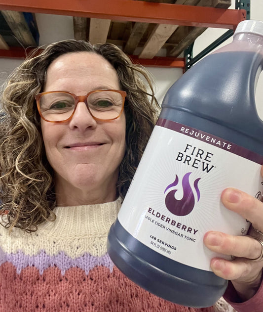 Woman in colorful knit sweater holding a large bottle of Fire Brew Elderberry apple cider vinegar tonic, smiling.