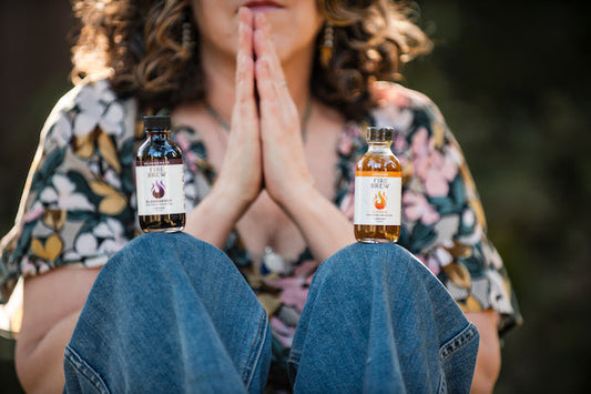 Woman seated in a prayer pose with two Fire Brew bottles balanced on her knees outdoors.