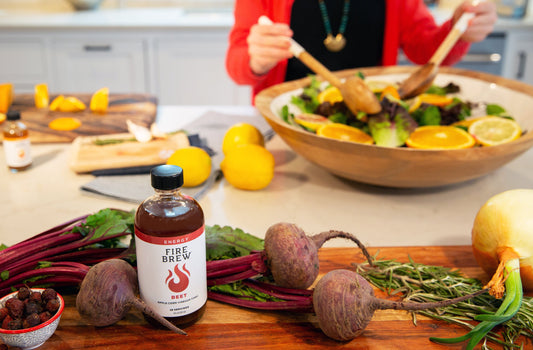 Bottle of Fire Brew Beet tonic on a kitchen counter surrounded by fresh beets, herbs, and salad ingredients.
