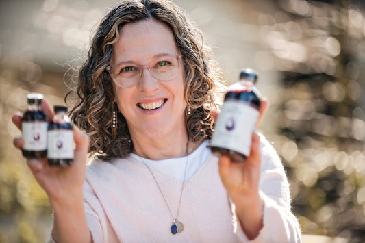 Smiling woman outdoors holding three bottles of Fire Brew herbal tonic in both hands.