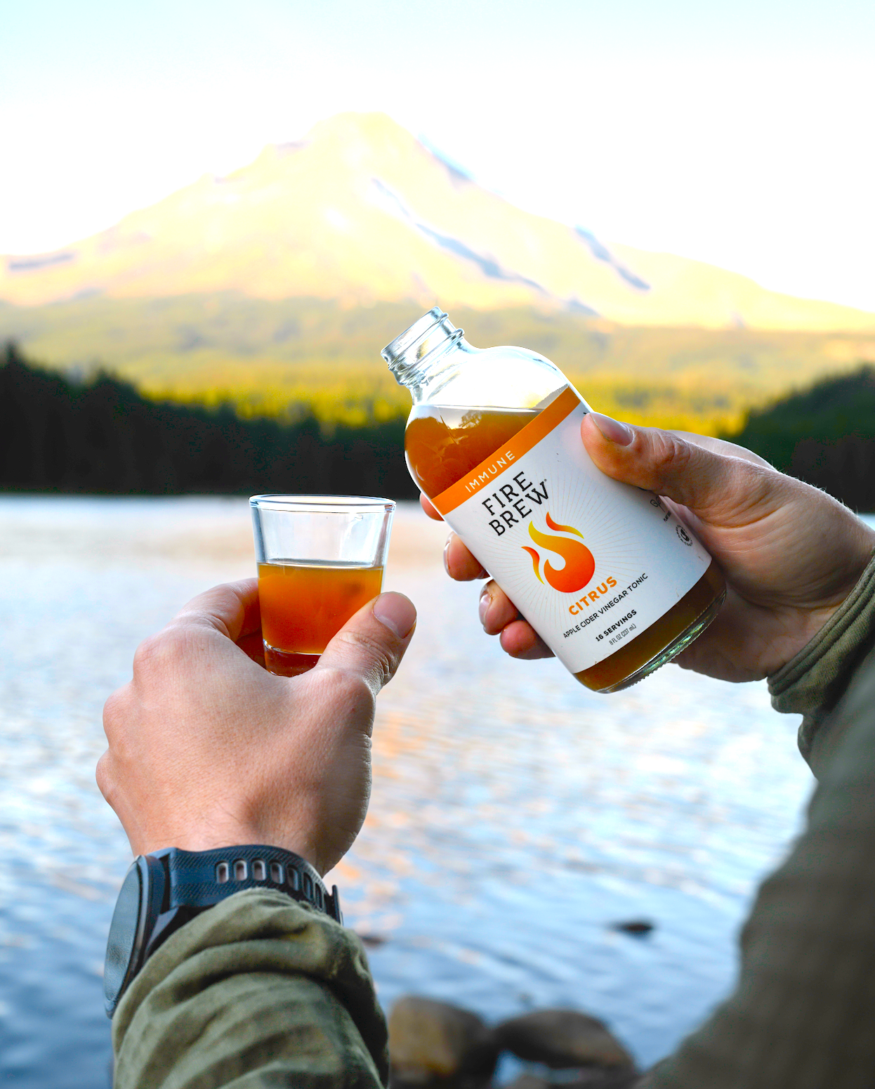 Person pours Fire Brew Citrus Apple Cider Vinegar tonic into shot glass by lake with mountain in background.