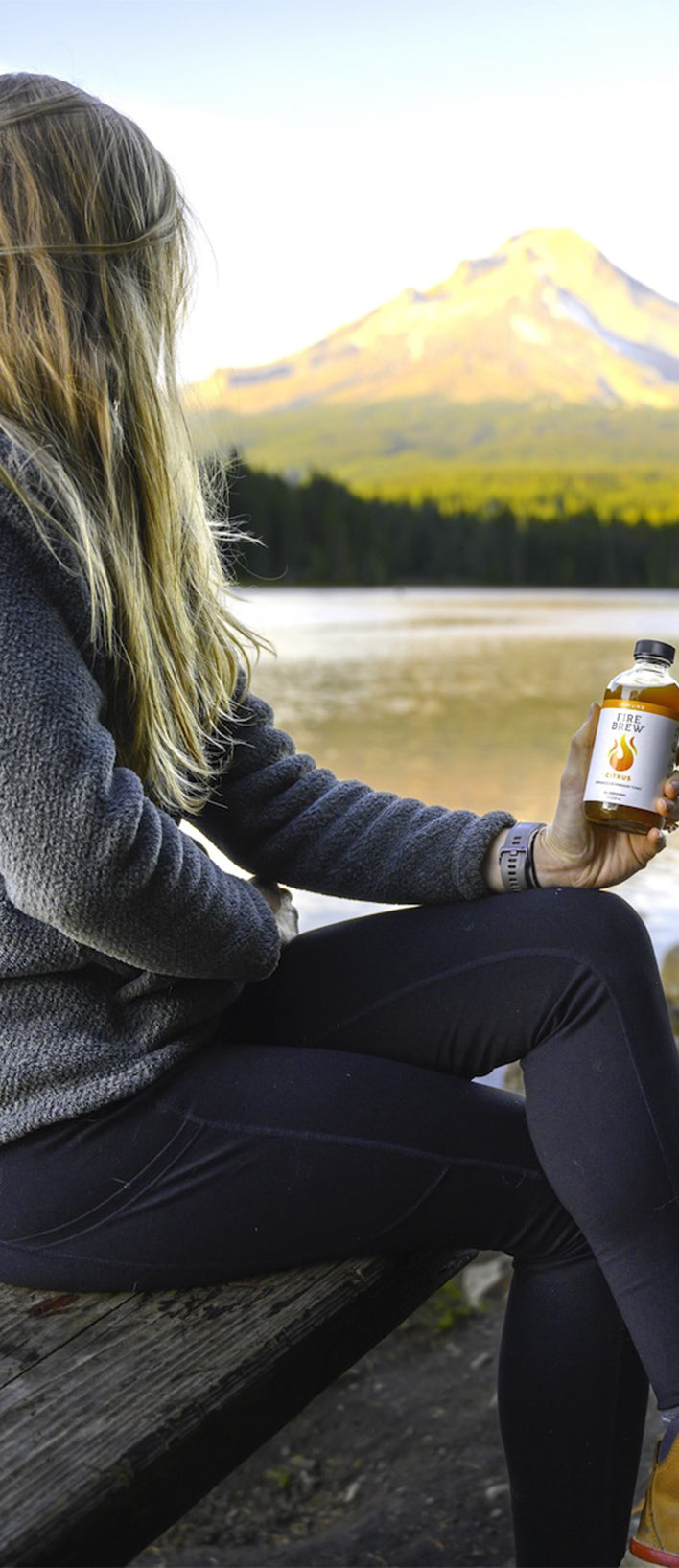 Woman on a bench near lake and mountains holding Fire Brew Citrus apple cider vinegar tonic bottle.