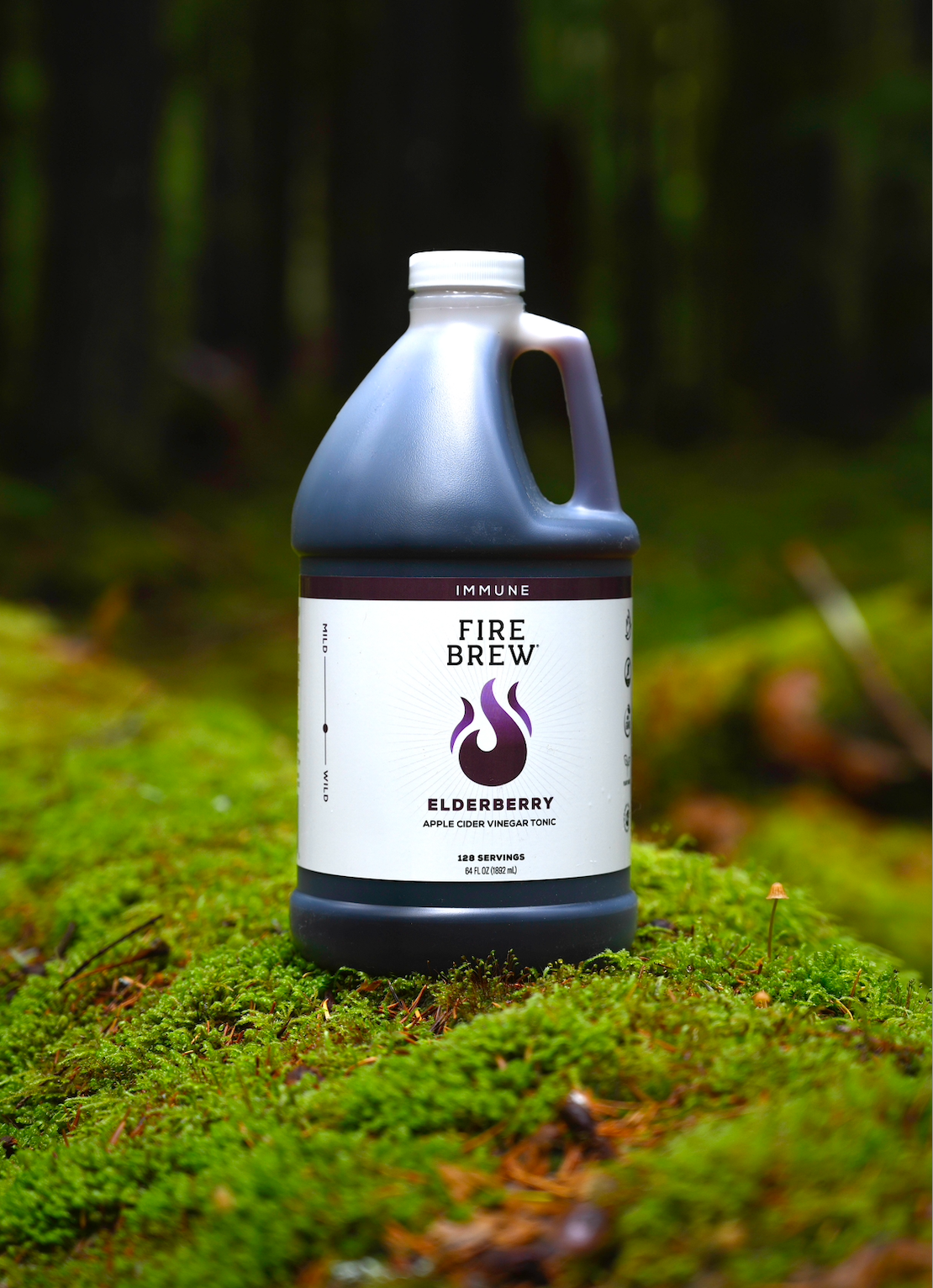 A bottle of Fire Brew Elderberry Apple Cider Vinegar Tonic stands on a lush green moss-covered forest floor.