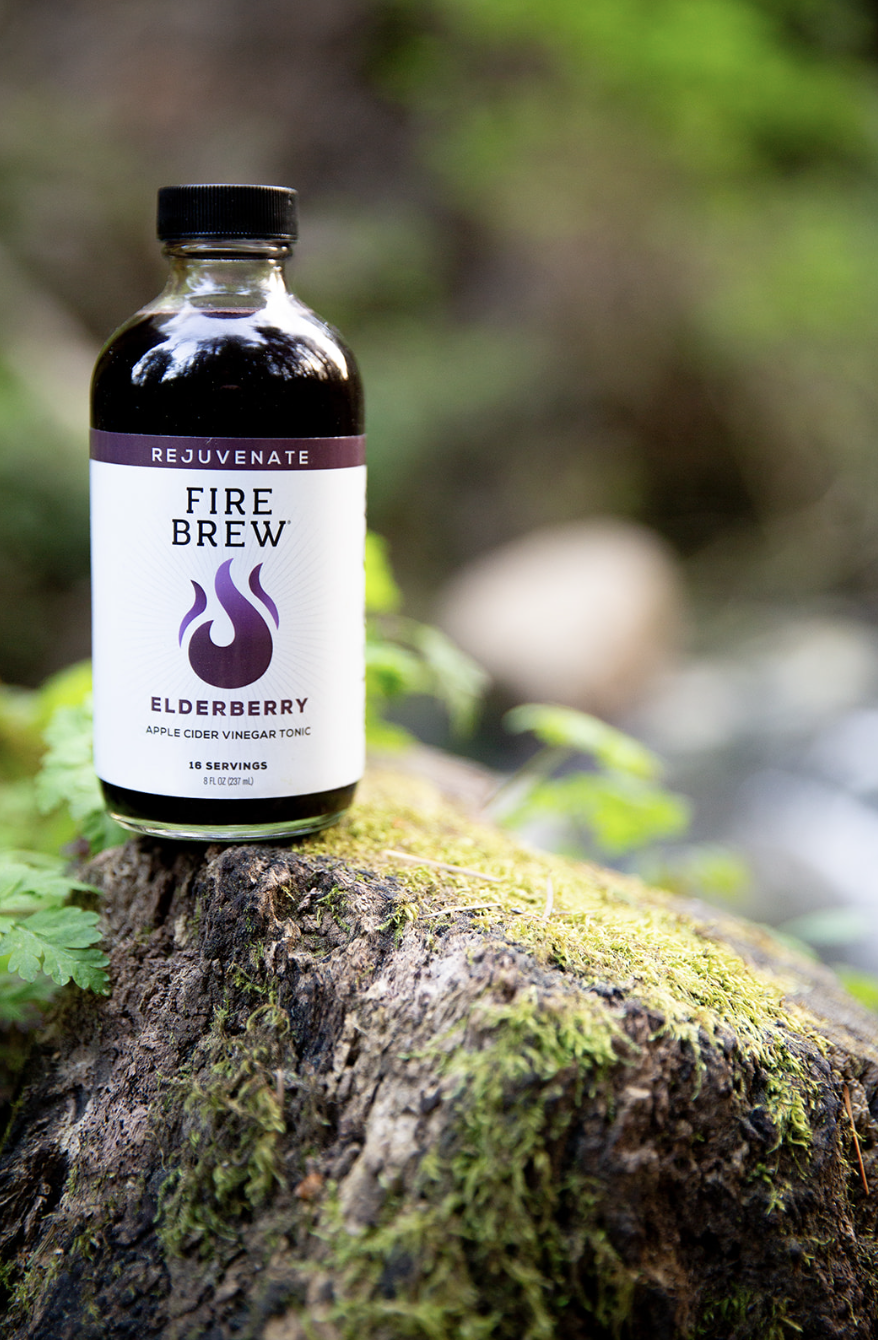 Bottle of Fire Brew Elderberry apple cider vinegar tonic placed on mossy tree stump in nature setting.