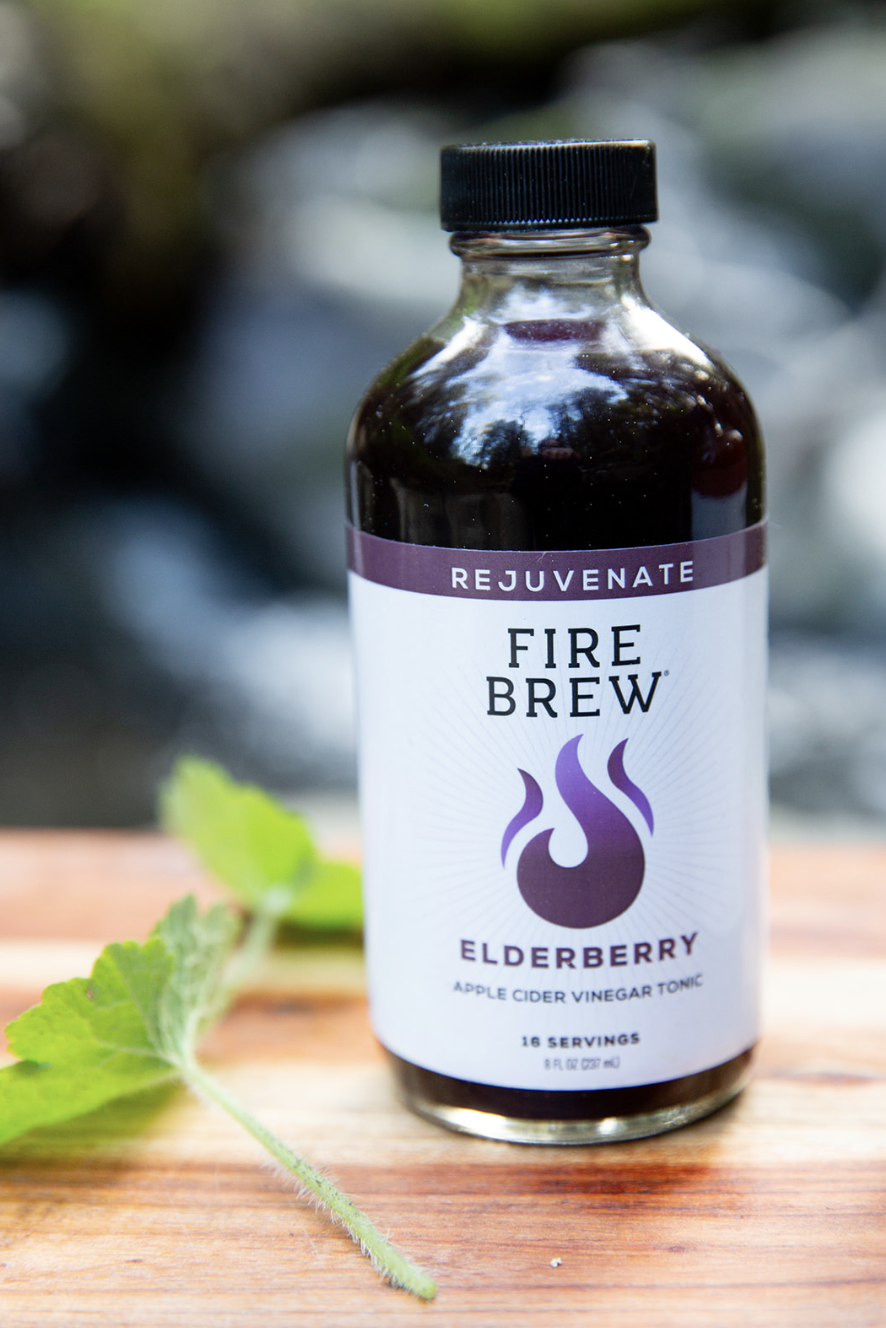 A bottle of Fire Brew elderberry apple cider vinegar tonic sits on a wooden surface beside herbs.