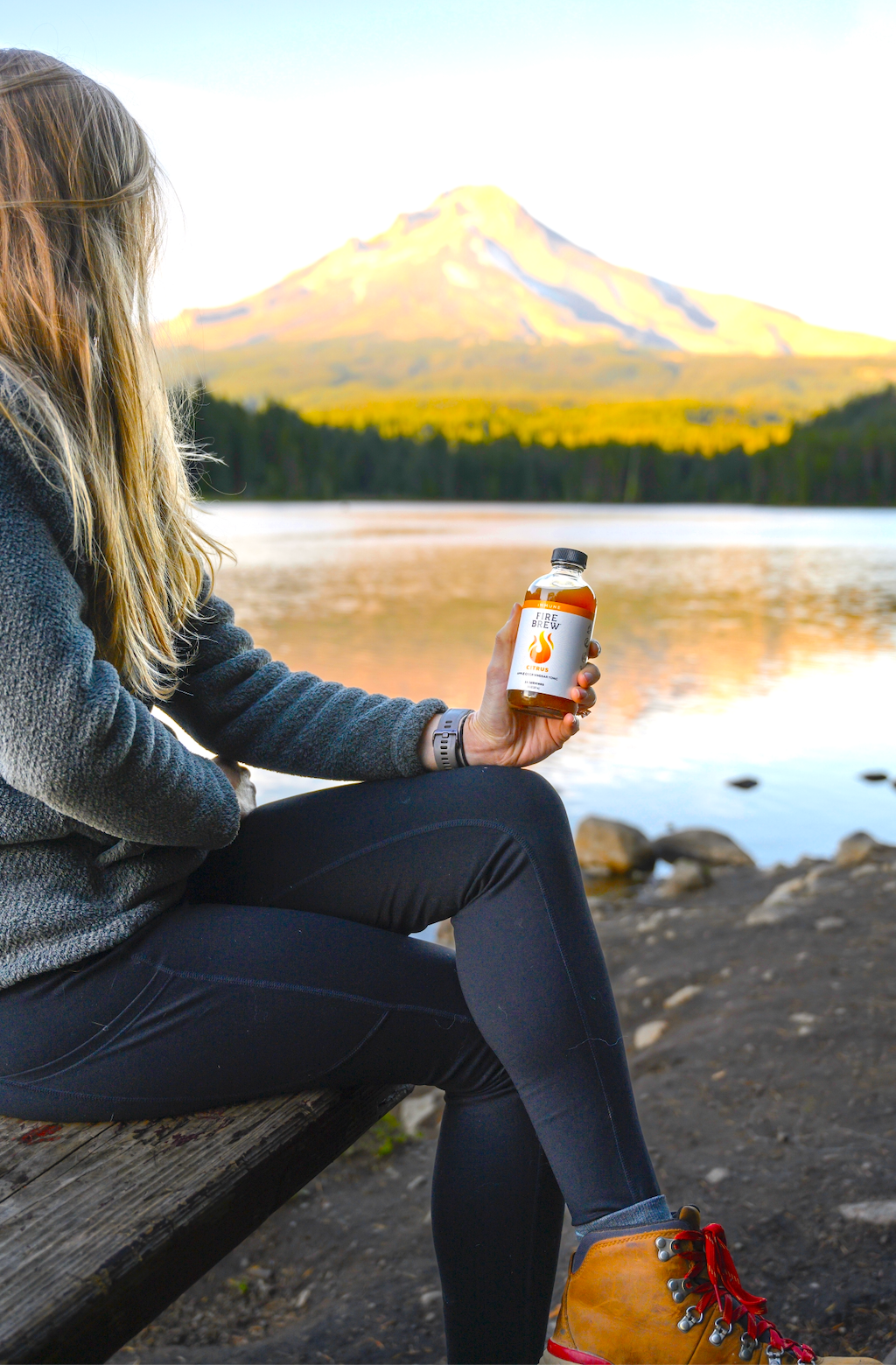 A woman holds a Fire Brew Citrus Apple Cider Vinegar tonic while sitting lakeside with a mountain view.