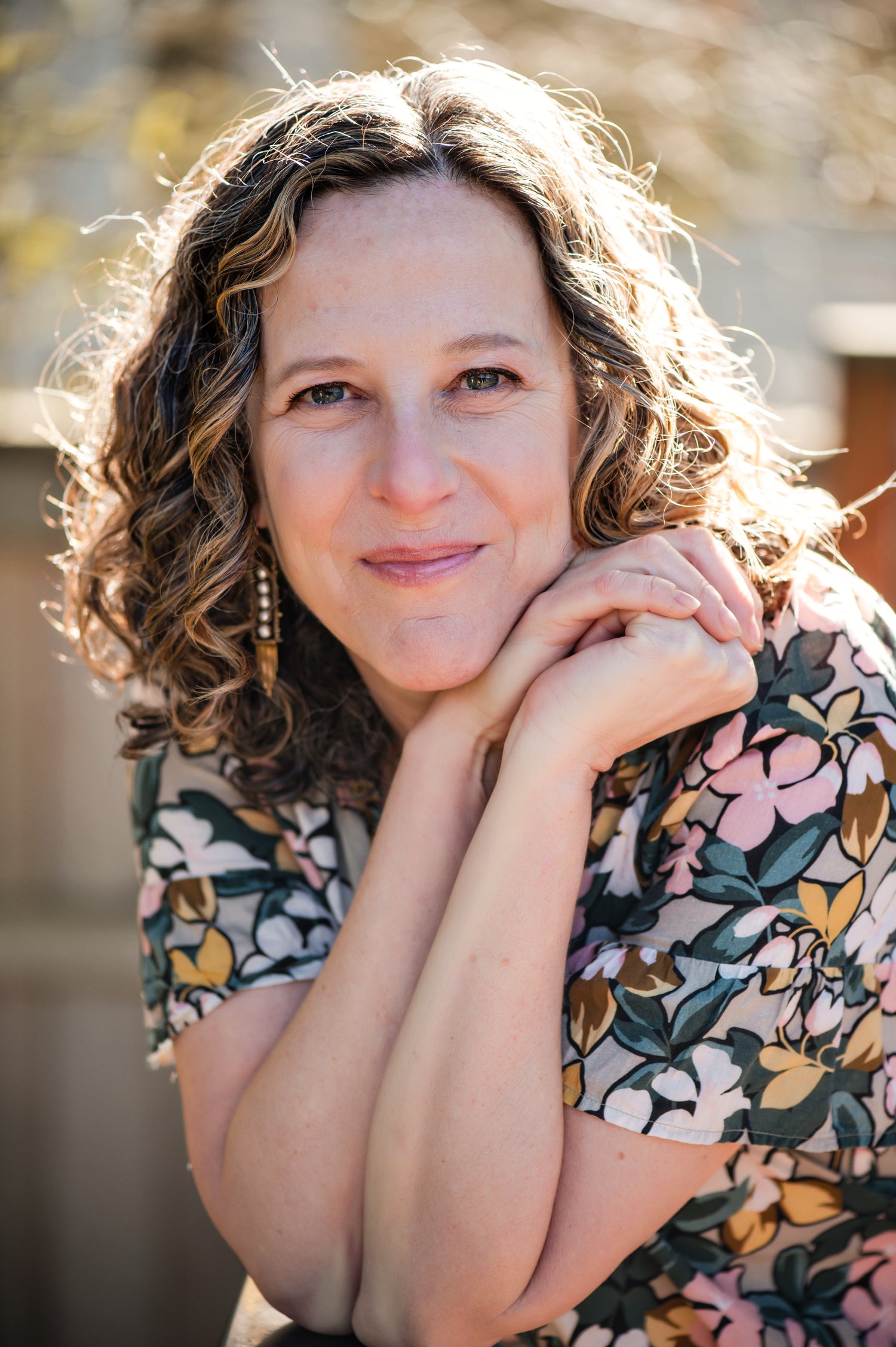 Woman with curly hair and floral shirt smiles warmly, resting chin on hands in sunlight.