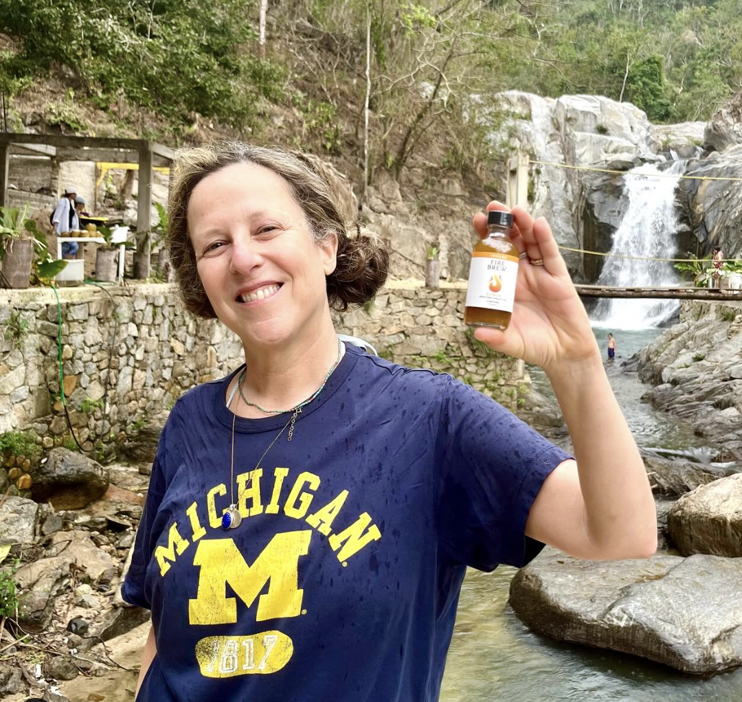 Smiling woman in Michigan shirt holds Fire Brew bottle by a rocky waterfall in a lush forest setting.