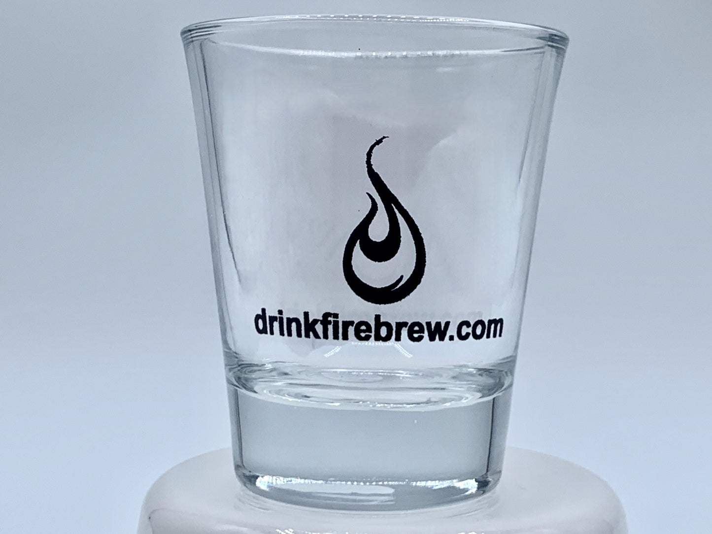 Clear shot glass with fire logo and "drinkfirebrew.com" printed in black on a white base.