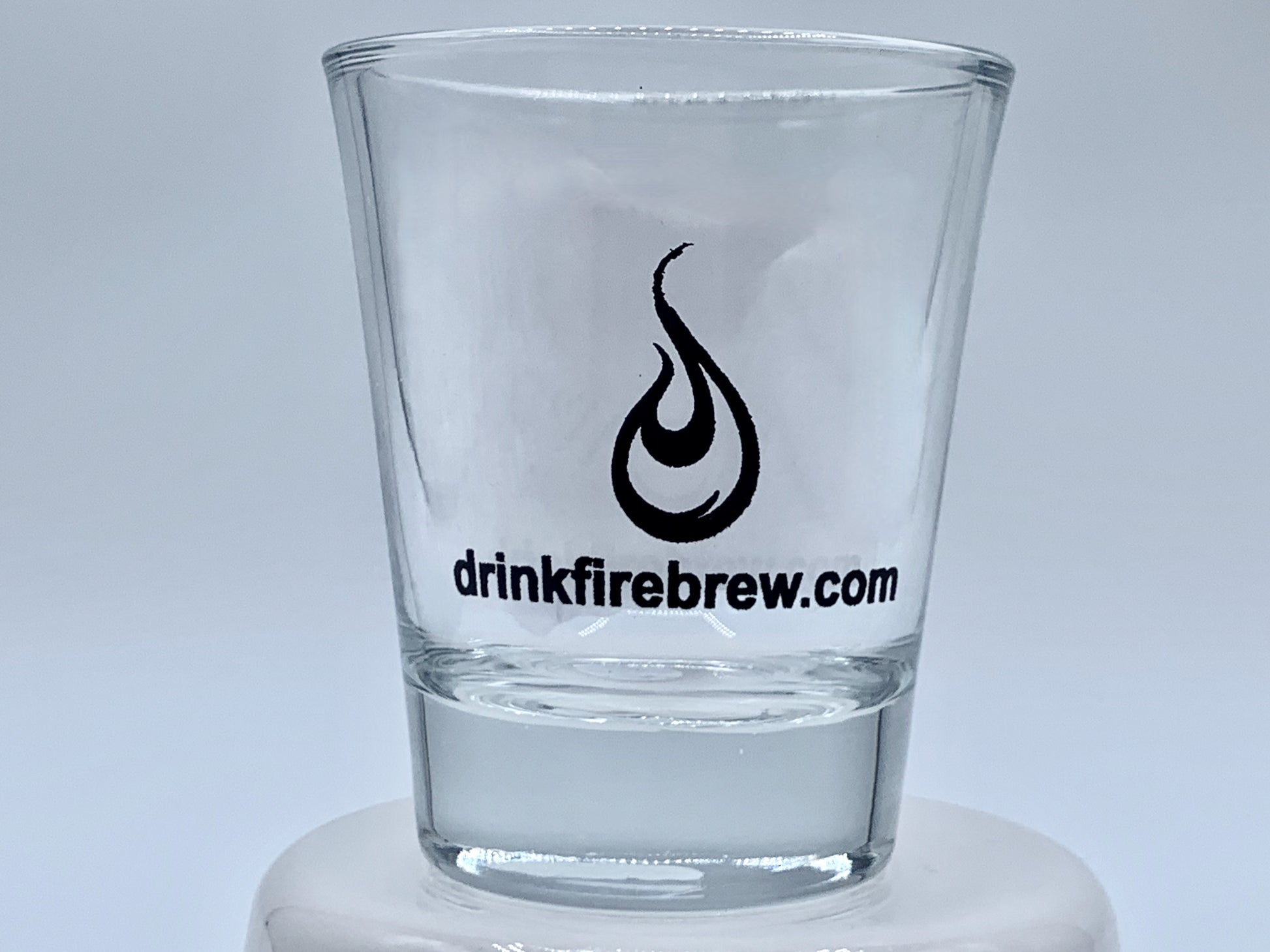 Clear shot glass with fire logo and "drinkfirebrew.com" printed in black on a white base.