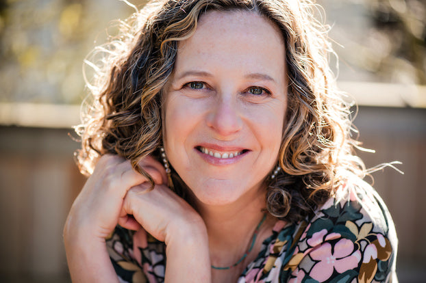 Smiling woman with curly hair and floral top sitting outdoors in soft natural sunlight.