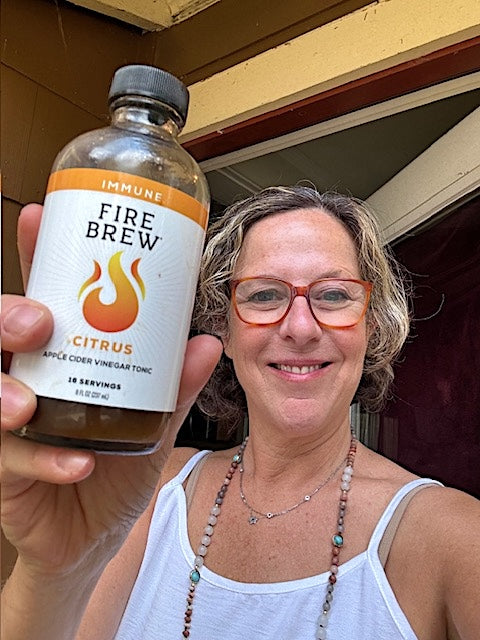 Smiling woman holding a bottle of Fire Brew citrus immune tonic with apple cider vinegar in front of her home.