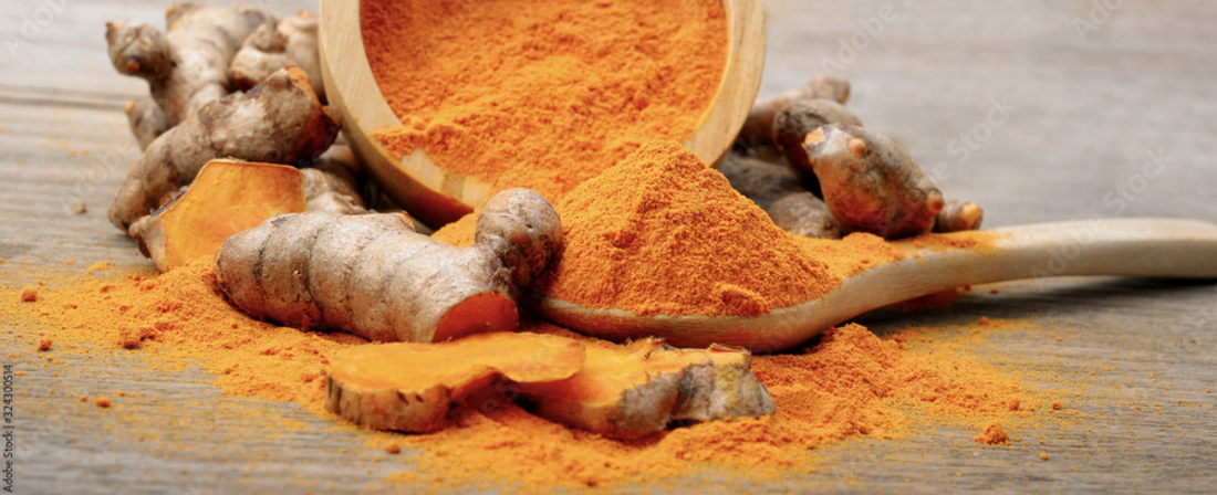Fresh turmeric roots and slices next to a wooden spoon and bowl filled with vibrant orange turmeric powder.